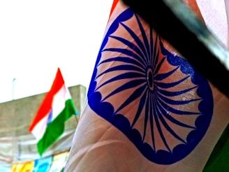 A close-up view of a fabric featuring a blue wheel design known as the Ashoka Chakra. In the blurred background, there is a tricolor flag with orange, white, and green horizontal stripes.