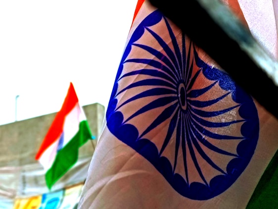 A close-up view of a fabric featuring a blue wheel design known as the Ashoka Chakra. In the blurred background, there is a tricolor flag with orange, white, and green horizontal stripes.