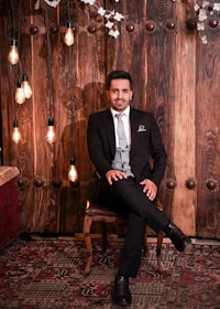 A man in a black suit and tie sits on a chair against a background of rustic, wooden panels. The setting includes hanging Edison-style light bulbs and decorative flowers. A richly patterned carpet covers the floor, adding to the warm and elegant ambiance.