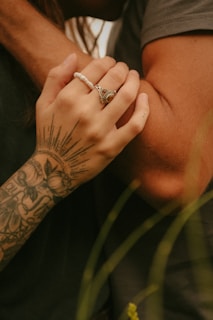 A close-up of two arms, one with a tattoo and a beaded bracelet, gently holding the other. The tattoo features intricate designs and floral motifs, while the bracelet is white with a large ornate ring on the finger. The background and lighting suggest an intimate, serene setting.