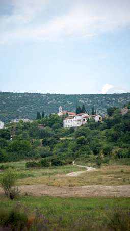 A scenic landscape featuring a small church with a steeple located amidst lush greenery on a hill. The building has a red-tiled roof and pale walls, surrounded by trees and bushes. A dirt path meanders through the grassy foreground, leading toward the hill. The sky above is partly cloudy with blue tones.
