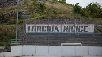 A hillside sports area with a concrete wall displaying the text 'Torcida Ricice' in bold letters. The surroundings include greenery and rocks, suggesting a natural backdrop. The area seems to be an outdoor sports field with metal fencing and a white, weathered soccer goal.
