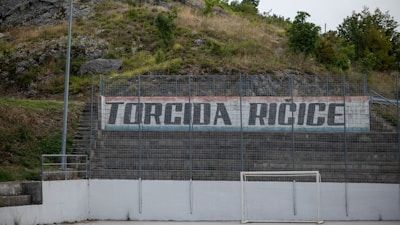 A hillside sports area with a concrete wall displaying the text 'Torcida Ricice' in bold letters. The surroundings include greenery and rocks, suggesting a natural backdrop. The area seems to be an outdoor sports field with metal fencing and a white, weathered soccer goal.