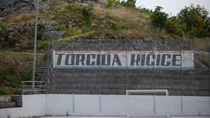 A hillside sports area with a concrete wall displaying the text 'Torcida Ricice' in bold letters. The surroundings include greenery and rocks, suggesting a natural backdrop. The area seems to be an outdoor sports field with metal fencing and a white, weathered soccer goal.