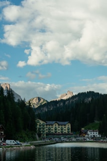 A picturesque landscape features a serene lake in the foreground, reflecting the surrounding scenery. Behind the lake, there is a charming multi-story building with green accents, likely a hotel or lodge. Tall evergreen trees frame the scene, and distant mountains with rocky peaks rise against a backdrop of a partly cloudy sky. The environment suggests a tranquil, natural setting.