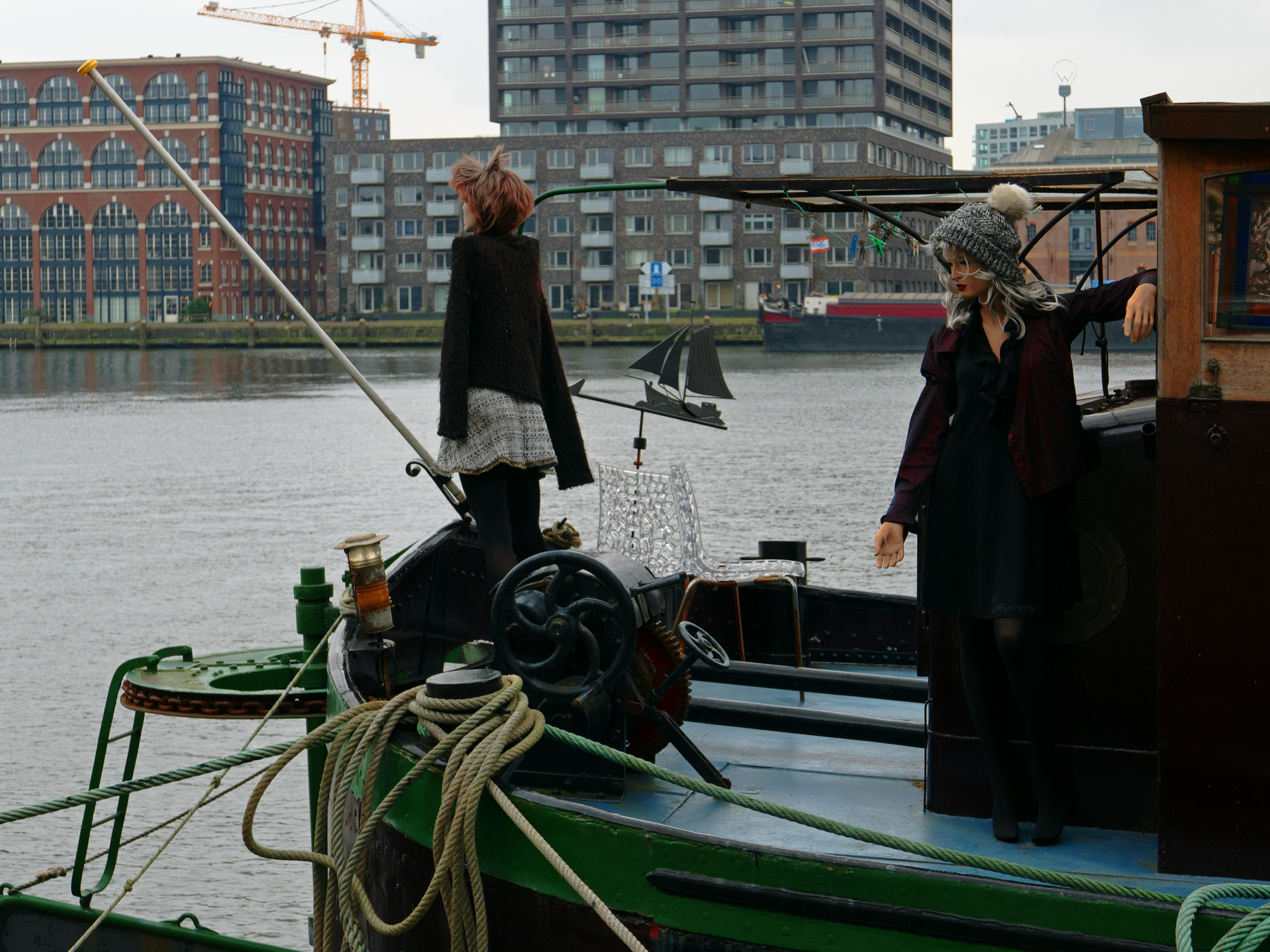 two people standing on a boat in the water, A houseboat, was a former transport ship but now people are living here for many years already, together with their dolls and puppets, standing and sitting on the deck Location is the harbor along the Javakade in Amsterdam city with its grey water and light. In the background facades of residential buildings, built along the water border.