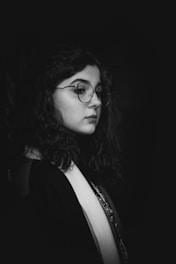 A black and white portrait of a person with wavy dark hair and glasses, gazing thoughtfully into the distance. The lighting creates a high contrast, highlighting the contours and features of their face.