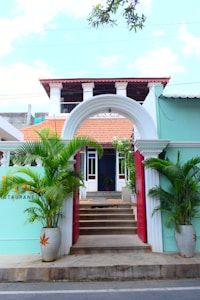 A vibrant, inviting entrance features a white arched gateway with red doors leading to a staircase. The building has a teal facade with red-tiled roofing and white pillars. Lush green potted plants flank the entrance, and a partially visible sign suggests it's a restaurant.