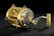 A metallic fishing reel with a gold finish is covered in water droplets, reflecting light on its shiny surface. It is placed against a dark background that highlights its intricate design and craftsmanship.