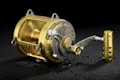 A metallic fishing reel with a gold finish is covered in water droplets, reflecting light on its shiny surface. It is placed against a dark background that highlights its intricate design and craftsmanship.