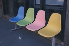 Four colorful plastic chairs are aligned in a row outside a building. Each chair is a different color: blue, green, pink, and yellow. They are mounted on a shared metal base. The chairs are placed on a concrete surface near a large window.
