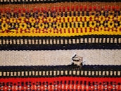 A woven textile featuring multiple horizontal stripes with intricate patterns. The colors include red, yellow, brown, black, and cream, each forming distinct geometric motifs. A small tear is present in the middle of a beige stripe.