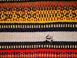 A woven textile featuring multiple horizontal stripes with intricate patterns. The colors include red, yellow, brown, black, and cream, each forming distinct geometric motifs. A small tear is present in the middle of a beige stripe.