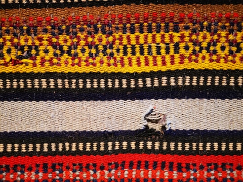 A woven textile featuring multiple horizontal stripes with intricate patterns. The colors include red, yellow, brown, black, and cream, each forming distinct geometric motifs. A small tear is present in the middle of a beige stripe.