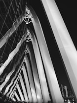 A black and white architectural photograph featuring large, curved, illuminated beams extending into the night sky. The beams form an arching pattern, with reflective glass structures on the left, showing mirrored patterns. The city skyline can be seen in the background with a few tall buildings.
