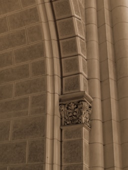 A close-up of a stone architectural element featuring a series of textured rectangular blocks. The central focus is on an intricately carved capital with floral and leaf designs, highlighting the craftsmanship. The image is captured in a neutral sepia tone that emphasizes the details of the stonework.