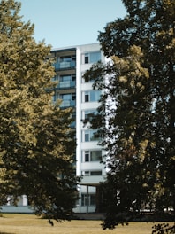 A mid-rise residential building with multiple balconies is partially obscured by lush green trees. The building's facade is white with a modern design, and it stands within a landscape of grass and foliage.