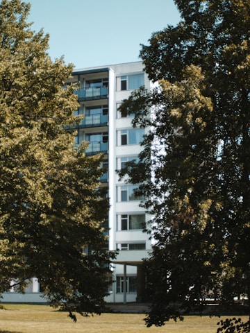 A mid-rise residential building with multiple balconies is partially obscured by lush green trees. The building's facade is white with a modern design, and it stands within a landscape of grass and foliage.