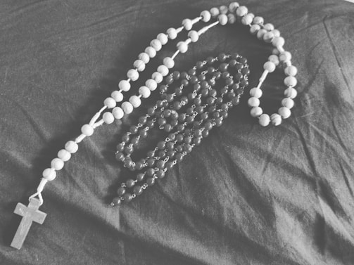 A monochrome photograph of two rosaries arranged on a dark fabric background. One rosary has larger wooden beads and is positioned in a loose circular pattern, while the other has smaller beads with a more intricate design.