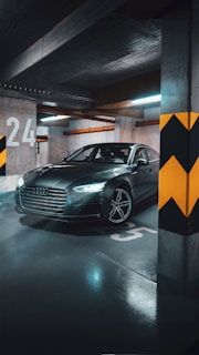 A sleek, modern car is parked in an underground parking garage. The vehicle is positioned near a concrete pillar with yellow and black chevron markings. The garage floor reflects the overhead lighting, creating a moody atmosphere. Large, white numbers are painted on the ground and walls.