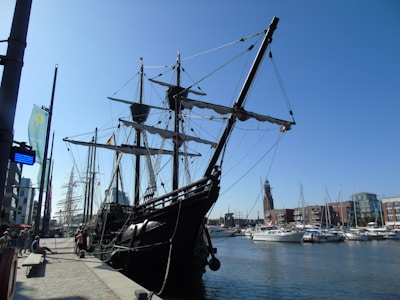 A large, old-fashioned sailing ship docked at a marina, with intricate rigging and tall masts. The background includes modern buildings, smaller boats, and a lighthouse-like structure. The sky is clear and blue, creating a bright atmosphere.