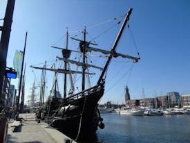 A large, old-fashioned sailing ship docked at a marina, with intricate rigging and tall masts. The background includes modern buildings, smaller boats, and a lighthouse-like structure. The sky is clear and blue, creating a bright atmosphere.