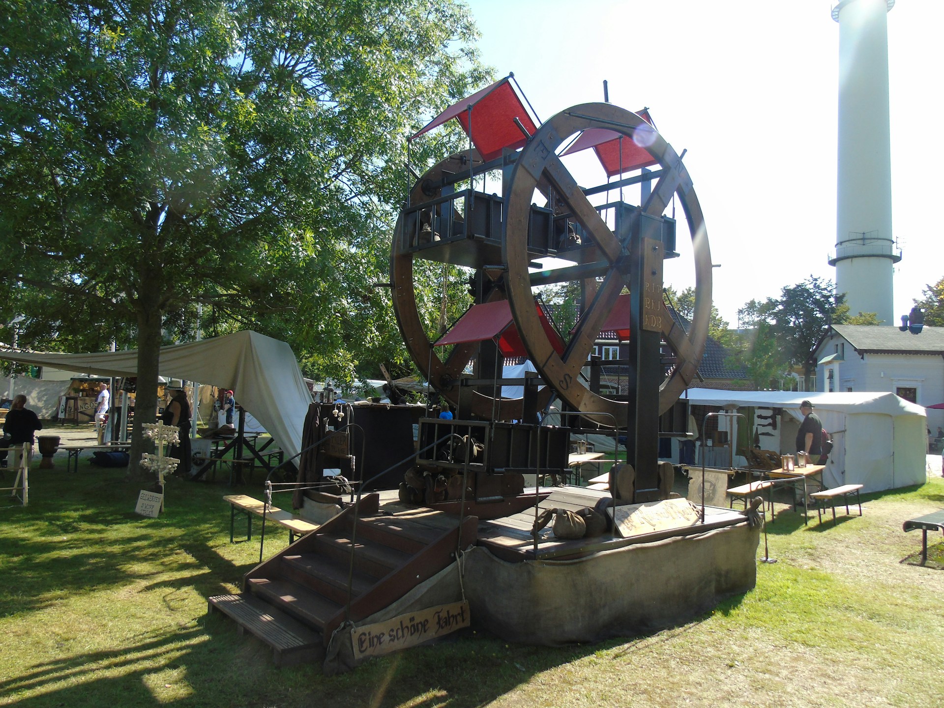 A large, rustic Ferris wheel-like structure is set up outdoors, surrounded by trees and tents, with signs in what appears to be German. People are milling around the area, and the scene is well-lit with sunlight casting shadows on the grass.