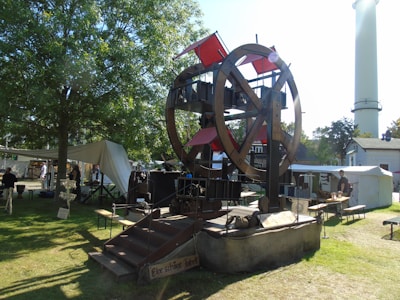 A large, rustic Ferris wheel-like structure is set up outdoors, surrounded by trees and tents, with signs in what appears to be German. People are milling around the area, and the scene is well-lit with sunlight casting shadows on the grass.