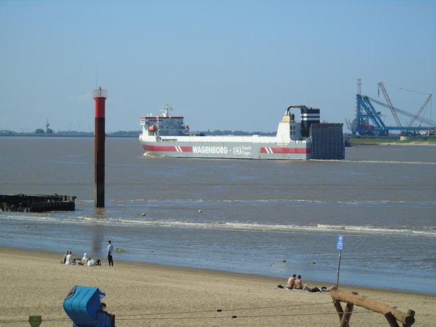 A large cargo ship with the name 'WAGENBORG' is navigating through a wide river or estuary. The ship is white with red accents and appears to belong to the company Saudi Kappa as indicated on its side. In the foreground, a sandy beach is visible with several people sitting and relaxing. A red and black navigation marker stands in the water near the shore. In the background, there are industrial structures and equipment, possibly part of a port or dock.