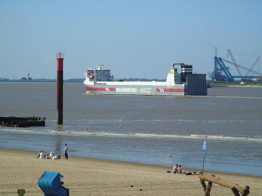 A large cargo ship with the name 'WAGENBORG' is navigating through a wide river or estuary. The ship is white with red accents and appears to belong to the company Saudi Kappa as indicated on its side. In the foreground, a sandy beach is visible with several people sitting and relaxing. A red and black navigation marker stands in the water near the shore. In the background, there are industrial structures and equipment, possibly part of a port or dock.