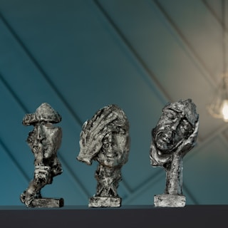 Three abstract sculptures of human heads displayed on a dark surface. Each sculpture appears textured and rustic with expressive facial features. The background is a soft blue with diagonal lines and a warm light illuminating part of the scene.