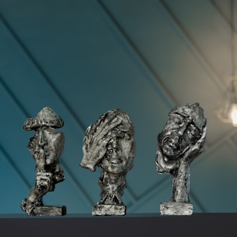 Three abstract sculptures of human heads displayed on a dark surface. Each sculpture appears textured and rustic with expressive facial features. The background is a soft blue with diagonal lines and a warm light illuminating part of the scene.