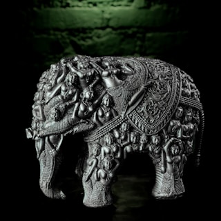 A detailed silver sculpture of an elephant adorned with intricate carvings of numerous human figures and ornate patterns. The craftsmanship displays a high level of detail with each figure appearing to be in various dynamic poses, contributing to the overall texture of the elephant. The backdrop is a dark greenish hue that highlights the metallic sheen of the sculpture.