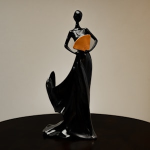 A sleek black statue of a stylized human figure standing with an elegant pose. The figure is holding an orange fan close to the body, and the lower part of the statue flows like a gown, creating a sense of movement.