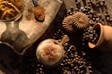 A cup of frothy coffee topped with cinnamon, surrounded by coffee beans and cookies placed on a textured burlap surface. A wooden spoon with ground coffee and a clay pot add rustic elements to the setting, illuminated by warm, natural light.