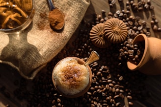 A cup of frothy coffee topped with cinnamon, surrounded by coffee beans and cookies placed on a textured burlap surface. A wooden spoon with ground coffee and a clay pot add rustic elements to the setting, illuminated by warm, natural light.