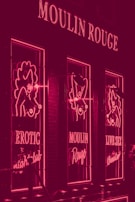 A neon-lit facade featuring signs and images related to a nightclub. The lights are predominantly pink, with the words 'Moulin Rouge', 'Erotic Nightclub', and 'Live Sex Theatre' visible. The design includes stylized figures outlined in neon.
