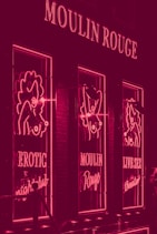A neon-lit facade featuring signs and images related to a nightclub. The lights are predominantly pink, with the words 'Moulin Rouge', 'Erotic Nightclub', and 'Live Sex Theatre' visible. The design includes stylized figures outlined in neon.