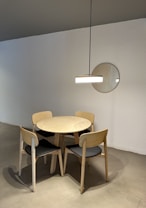 A minimalist room features a round wooden table surrounded by four wooden chairs with black seats. A modern pendant light hangs overhead, casting subtle shadows. A round mirror is mounted on the white wall.