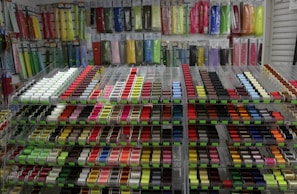 A wide array of colorful ribbons and threads are neatly organized on shelves. The ribbons are displayed in vertical packages hanging on the wall, and the threads are arranged in rows on spools. The scene is bright and vibrant with a diverse spectrum of colors ranging from reds and pinks to blues and greens.