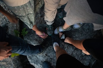 A group of people with various footwear, including sandals, sneakers, and crocs, standing on a rocky surface. The legs and feet are the focus, with different styles and colors of shoes contrasted against the gray texture of the rocks.