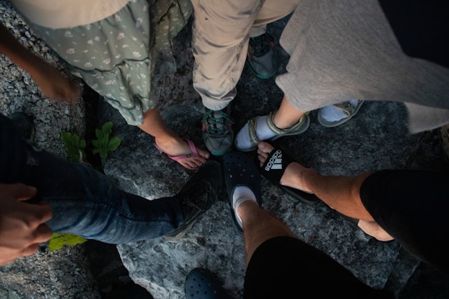 A group of people with various footwear, including sandals, sneakers, and crocs, standing on a rocky surface. The legs and feet are the focus, with different styles and colors of shoes contrasted against the gray texture of the rocks.