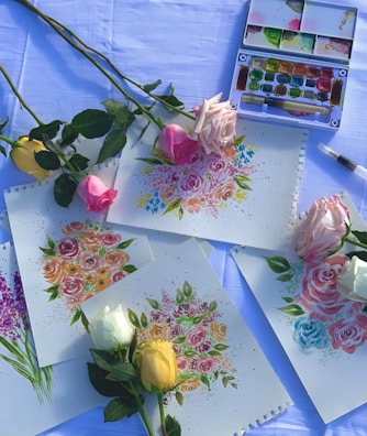 Multiple paintings depicting colorful floral arrangements are spread across a surface that appears to be covered with a light blue cloth. Real roses in various colors, including yellow, pink, and white, are placed on top of and around the paintings. There is a watercolor paint set and a brush visible in the background.