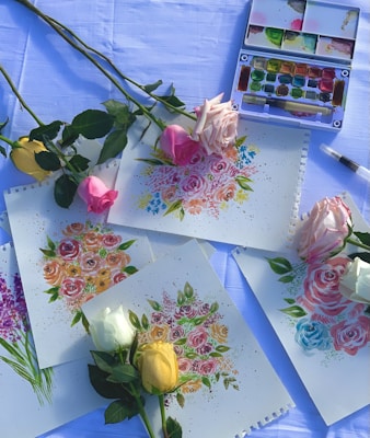 Multiple paintings depicting colorful floral arrangements are spread across a surface that appears to be covered with a light blue cloth. Real roses in various colors, including yellow, pink, and white, are placed on top of and around the paintings. There is a watercolor paint set and a brush visible in the background.