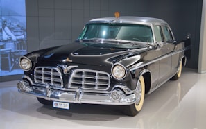 A classic vintage car with a polished black exterior and chrome detailing stands in an indoor showroom. The car has a distinctive front grille, round headlights, and white-walled tires, evoking a sense of elegance and luxury.