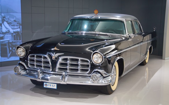 A classic vintage car with a polished black exterior and chrome detailing stands in an indoor showroom. The car has a distinctive front grille, round headlights, and white-walled tires, evoking a sense of elegance and luxury.