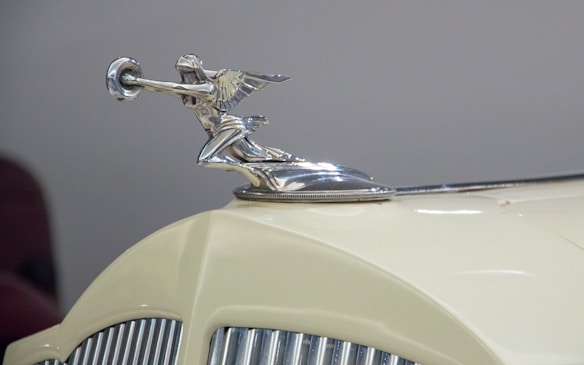 A chrome hood ornament shaped like a winged figure mounted on a vintage car with a cream-colored body. The car grille is visible, featuring vertical chrome bars.