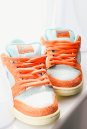 A pair of sneakers with a vibrant color scheme featuring orange suede sections and light teal fabric with orange laces. The shoes have labels on the tongue displaying the brand logo and are placed next to a white curtain, suggesting they are exposed to natural light, which highlights their colors.