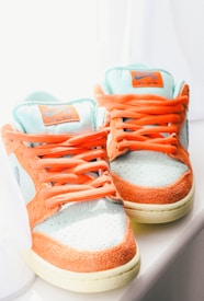 A pair of sneakers with a vibrant color scheme featuring orange suede sections and light teal fabric with orange laces. The shoes have labels on the tongue displaying the brand logo and are placed next to a white curtain, suggesting they are exposed to natural light, which highlights their colors.