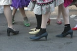 A group of people are standing closely together, wearing colorful and varied footwear, including high heels and socks. Skirts with different patterns and textures are visible, adding a playful and festive atmosphere.